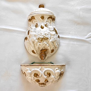 May include: A white ceramic wall fountain with gold-painted accents, featuring relief carvings of cherubs and scrolling foliage. The set includes a top reservoir with a central spout and a matching semi-circular basin with a green wire hanger.