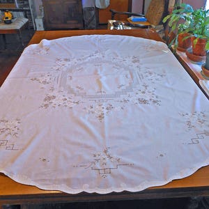 May include: A white tablecloth with embroidered floral and geometric patterns. The tablecloth is spread across a wooden table, showcasing the detailed stitching and scalloped edges. The embroidery features a combination of floral and geometric designs.