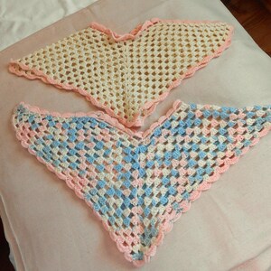May include: Two triangular crochet shawls featuring openwork patterns. The top shawl is cream-colored with a light pink scalloped border. The bottom shawl features a multicolored pattern of light blue, cream, and light pink with a pink scalloped edge.