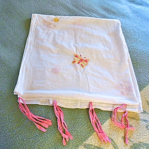 May include: White fabric with a butterfly embroidery and pink tassels. The fabric has a delicate appearance, with a butterfly design in the center and small pink accents. Pink tassels hang from the bottom edge, adding a decorative touch.