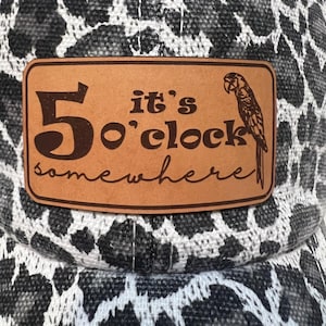Puede incluir: Gorra de béisbol con un estampado de animales en blanco, negro y gris. Un parche de cuero marrón presenta el texto "5 o'clock somewhere" con una ilustración de loro. Un accesorio casual.