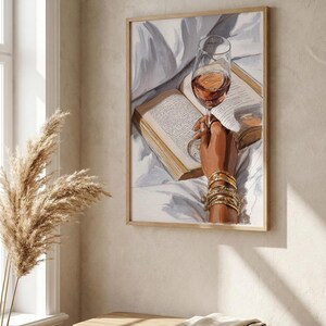 May include: A framed art print depicts a hand holding a wine glass over an open book. The hand is adorned with gold bracelets. The wine glass is filled with a rose-colored liquid. The artwork is set against a white background.