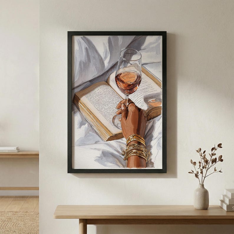 May include: Framed art print of a hand holding a wine glass filled with rose wine, resting on an open book. The hand is adorned with gold bracelets. The background features white bedding.