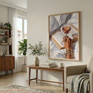 May include: A framed artwork depicts a hand holding a wine glass over an open book. The hand is adorned with gold bracelets. The artwork is in a room with a wooden bench, a vase with greenery, and a bookshelf.