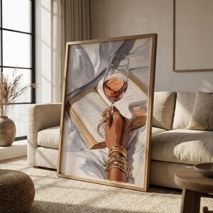 May include: A framed art print depicts a hand holding a glass of rose wine over an open book. The hand is adorned with gold bracelets. The artwork has a neutral color palette with soft lighting, creating a cozy and elegant atmosphere.