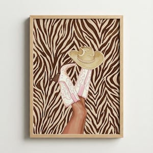 Op de afbeelding: Een ingelijst kunstwerk met een paar witte en roze cowboylaarzen en een bruine cowboyhoed. De laarzen hebben decoratieve stiksels en roze accenten. De achtergrond is een bruin- en tan zebraprint.