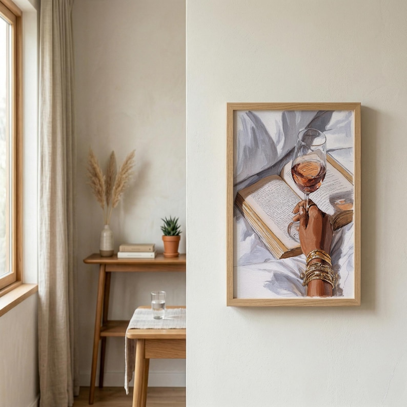 May include: A framed artwork depicts a hand holding a wine glass filled with rose wine, resting on an open book. The hand is adorned with gold bracelets. The artwork is in a light wood frame, suggesting a cozy, relaxing theme.