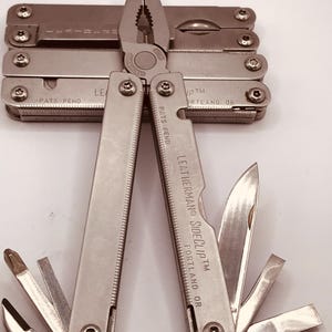 以下が含まれることがあります： プライヤー、ナイフ、その他のツールを備えたシルバーの Leatherman Sideclip マルチツール。 ツールは開いており、さまざまなコンポーネントが表示されています。 ハンドルには「LEATHERMAN SIDECUPTM PORTLAND OR」の文字が見えます。 他のマルチツールがいくつか後ろに積み重ねられています。