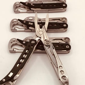 Könnte beinhalten: Eine Sammlung von Leatherman-Multitools mit silbernem und schwarzem Design. Die Werkzeuge umfassen Zangen, Klingen und andere Geräte. Das Leatherman-Logo ist sichtbar. Die Werkzeuge sind auf einer weißen Oberfläche angeordnet.