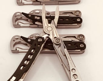 Könnte beinhalten: Eine Sammlung von Leatherman-Multitools mit silbernem und schwarzem Design. Die Werkzeuge umfassen Zangen, Klingen und andere Geräte. Das Leatherman-Logo ist sichtbar. Die Werkzeuge sind auf einer weißen Oberfläche angeordnet.