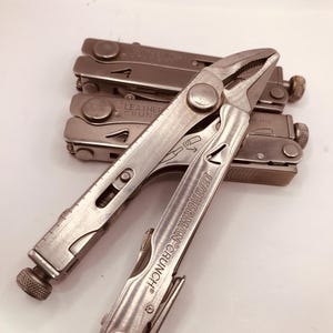 May include: A multi-tool with pliers, a knife, and other tools. The multi-tool is silver and has the text "LEATHERMAN CRUNCH" on the side. Several other multi-tools are stacked behind the main tool.