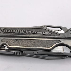Könnte beinhalten: Ein silbernes Leatherman Charge Multitool in geschlossener Position. Das Werkzeug verfügt über einen strukturierten Griff mit der Gravur LEATHERMAN Charge an der Seite. Eine Klinge mit der Aufschrift 154CM ist teilweise sichtbar. Das Gerät ist mit vier Sternschrauben befestigt.