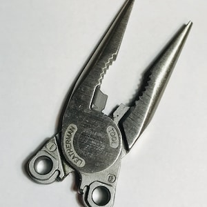 Könnte beinhalten: Ein silberfarbener Leatherman Tool Multifunktions-Zangenkopf mit spitz zulaufenden Backen, gezahnten Greifflächen und zwei kreisförmigen Drehpunkten an der Basis. Die Metalloberfläche ist mit dem Text LEATHERMAN TOOL geprägt.
