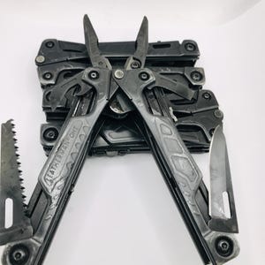 Può includere: Un utensile multifunzione Leatherman OHT nero con pinze spiegate, dotato di una lama seghettata sul manico sinistro e una lama a filo dritto sul manico destro. L'utensile ha una finitura opaca con rivetti e giunti meccanici visibili.