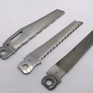 Könnte beinhalten: Drei Metallsägeblätter mit silbernem Finish vor weißem Hintergrund. Zwei Blätter haben gezahnte Kanten, eines hat eine feilenartige Oberfläche. Jedes Blatt hat ein Loch am Ende zur Befestigung.