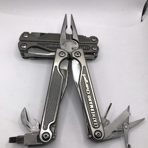 Pode incluir: Uma multi-ferramenta prateada e cinzenta com a marca "LEATHERMAN Charge®" visível. A ferramenta inclui alicates, uma faca, tesouras e outros implementos. Outra multi-ferramenta fechada está no fundo.