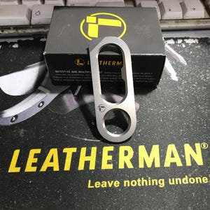 以下が含まれることがあります： 2 つの円形の開口部と長方形のトップを備えた、シルバーの Leatherman カラビナ。 Leatherman のロゴは、カラビナとパッケージに表示されます。 パッケージは黒で、黄と白のテキストが使用されています。
