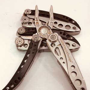 May include: A silver and black multi-tool with pliers and other implements. The brand name "LEATHERMAN Freestyle" is visible. Several similar tools are stacked in the background. The tools are made of metal and have a sleek design.