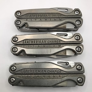 May include: Three Leatherman multi-tools are displayed, showcasing a silver-colored, rugged design. The top tool is labeled "LEATHERMAN TITANIUM," while the other two are marked "LEATHERMAN CHARGE." Each tool features multiple components and a compact, foldable design.