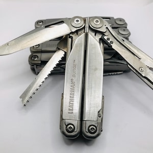 Könnte beinhalten: Ein silbernes Leatherman Surge Multifunktionswerkzeug mit mehreren ausgeklappten Klingen und Werkzeugen. Das Werkzeug hat eine gebürstete Metalloberfläche mit der Gravur LEATHERMAN SURGE auf dem Griff. Das Multifunktionswerkzeug ist in einem geöffneten, teilweise entfalteten Zustand dargestellt.