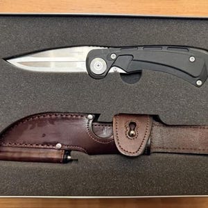 May include: A folding knife with a silver-toned blade and a black handle, positioned above a brown leather sheath with a snap closure. Both items are securely nestled within a rectangular grey foam insert inside a metal storage container.