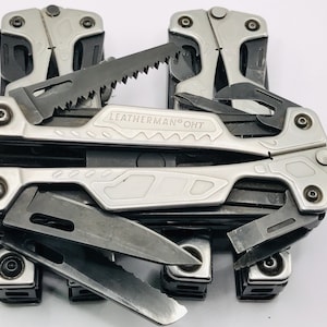 May include: A Leatherman OHT multi-tool featuring a silver-toned metal handle with the brand name and model engraved. The tool is partially unfolded, revealing several black-coated steel components including a saw blade, a knife, and a strap cutter.