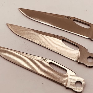 May include: Three stainless steel knife blades with a brushed finish. Each blade has a pointed tip and a hole near the handle. The text "S30V" is etched on each blade.