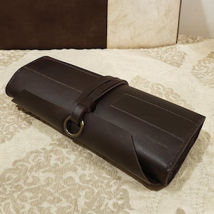 May include: A rolled-up, dark brown leather case with a strap and metal ring closure. The case is rectangular and appears to be made of a smooth, durable material. The design is simple and elegant, suitable for storing various items.