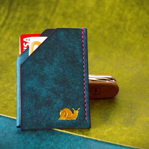 May include: Teal leather cardholder with a gold snail emblem and pink stitching. A red and white credit card is visible inside. A wooden Swiss Army knife rests beside the cardholder.