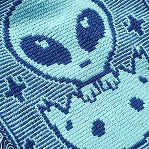 Pode incluir: Um cobertor de crochê feito à mão com um design alienígena pixelizado em tons de azul e verde-azulado. O cobertor tem uma borda com franjas e um padrão repetido de um rosto alienígena com olhos grandes e uma boca pequena. O tema geral é o espaço e extraterrestres.