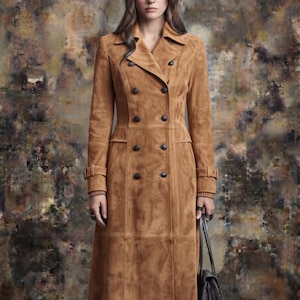 May include: A tan suede double-breasted trench coat featuring a wide notched collar, long sleeves with buckled cuffs, and two vertical rows of dark buttons. The coat extends to the mid-calf and has a structured fit with visible seam detailing.