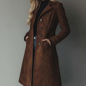 May include: A long, brown suede coat with a classic button-down design. The coat features a notched collar, long sleeves, and pockets. The coat is worn over a black turtleneck and blue jeans.