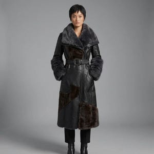 May include: A long black leather coat featuring a large grey faux fur collar, matching faux fur cuffs, and dark brown faux fur panels on the lower front. The coat includes a black leather belt with a buckle at the waist and a full-length front zipper.