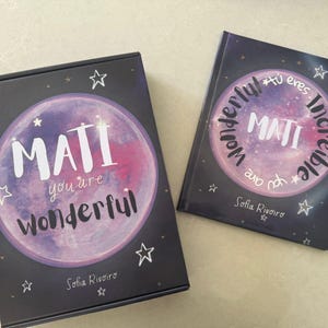 Könnte beinhalten: Zwei Artikel mit einem himmlischen Thema. Eines ist eine Box, das andere ein Buch, beide mit dunklem Hintergrund und einem zentralen Design mit einem rosa und lila Mond mit dem Namen "MATI" in Weiß. Auf der Box steht "you are wonderful".