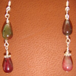 May include: A pair of dangle earrings featuring teardrop-shaped gemstones in shades of green, brown, and red. The stones are connected by silver-colored wire and hang from silver-colored ear wires. The earrings have a natural, earthy aesthetic.