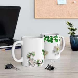 Puede incluir: Dos tazas de cerámica blancas con diseños florales. Una taza presenta flores de magnolia blancas y capullos verdes. La otra taza tiene hojas verdes y una flor blanca con un gatito asomando. Ambas tazas tienen un asa blanca.