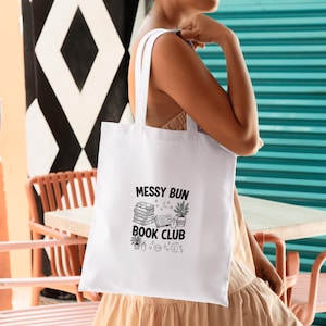 Könnte beinhalten: Weiße Tragetasche mit dem Text "Messy Bun Book Club" in Schwarz. Das Design umfasst Illustrationen von Büchern, Pflanzen und Himmelselementen. Die Tasche hat zwei weiße Griffe und wird von einer Person gehalten.