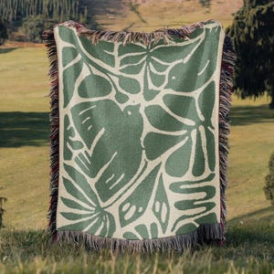 May include: A green and cream-colored woven throw blanket with a botanical design featuring leaves and a bird. The blanket has a fringed edge and is displayed outdoors on a grassy surface.