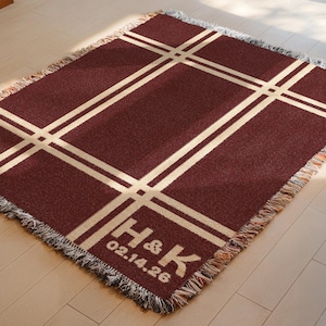 May include: A maroon throw blanket with a cream-colored grid pattern. The blanket has a fringed edge and features the initials "H & K" and the date "02.14.26" in the lower right corner. The blanket is rectangular and appears to be made of a soft material.