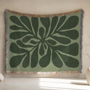 May include: A woven wall hanging with a large, abstract floral design in shades of green. The central motif is a dark green flower shape against a lighter green background. The edges have a fringed trim.