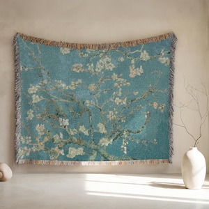 May include: A woven tapestry featuring a reproduction of Van Gogh's "Almond Blossoms" painting. The artwork depicts a blossoming almond tree against a teal background. The tapestry has a fringed edge and is suitable for wall decor.