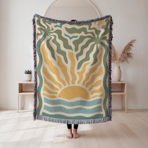 May include: Woven throw blanket featuring a stylized sunset design. The blanket showcases a yellow sun with radiating lines above blue waves. Palm tree silhouettes and a border in shades of green and blue complete the design. The blanket has fringed edges.