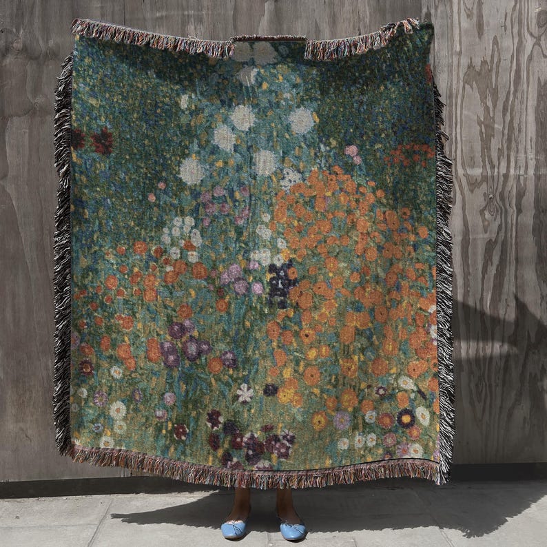 May include: A woven throw blanket featuring a floral design in shades of green, orange, white, and purple. The blanket has a fringed edge and is held up against a neutral background. The artwork depicts a field of flowers.