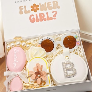 May include: A white gift box containing a pink hairbrush with the name Bnetehga, white sunglasses with Bnetehga lettering and flower accents, a white circular jewellery case with a pearl letter B, a hair clip, and a floral hair accessory.