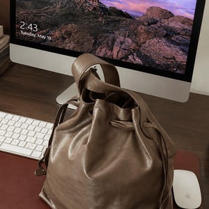 May include: A taupe-colored leather bucket bag featuring a drawstring closure and a single top handle. The bag has a smooth, slightly glossy finish and a soft, slouchy structure. The drawstring is threaded through metal eyelets at the top.