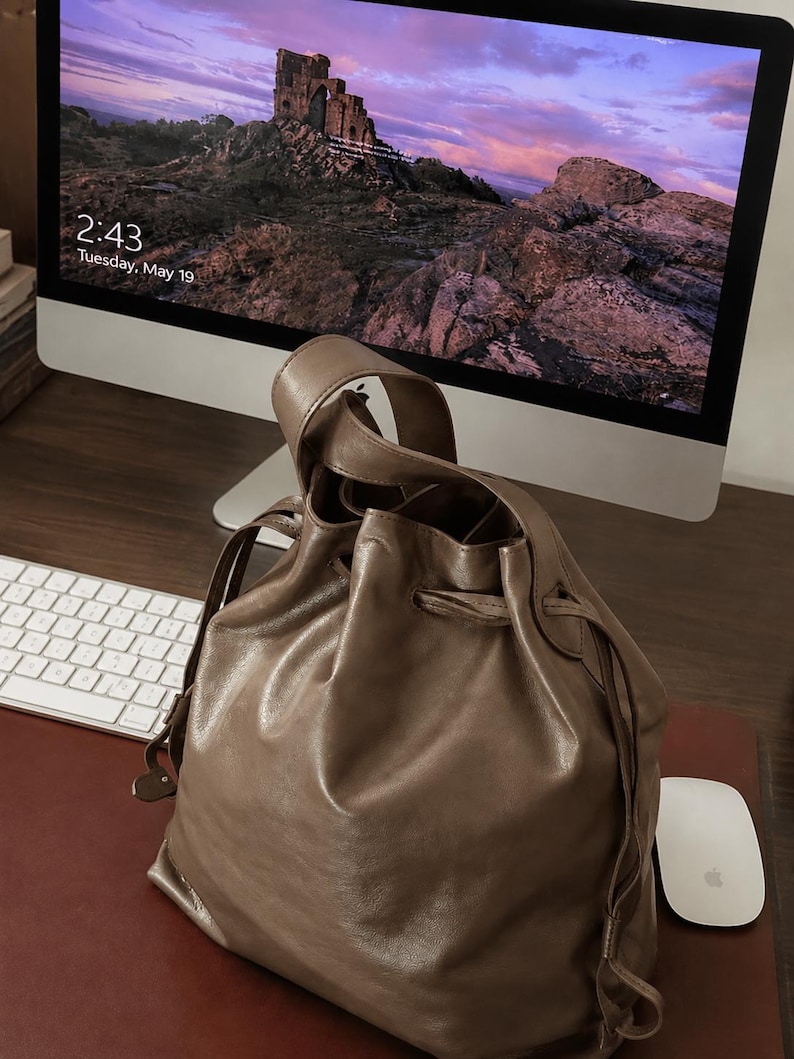 May include: A taupe-colored leather bucket bag featuring a drawstring closure and a single top handle. The bag has a smooth, slightly glossy finish and a soft, slouchy structure. The drawstring is threaded through metal eyelets at the top.