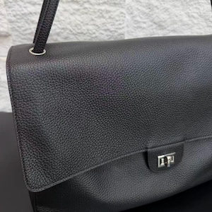Detailed front view of a black leather shoulder bag featuring a textured leather flap, silver turn-lock closure, and adjustable strap attachment. The minimalist luxury design combines timeless style with practical functionality for everyday outfits and occasions.