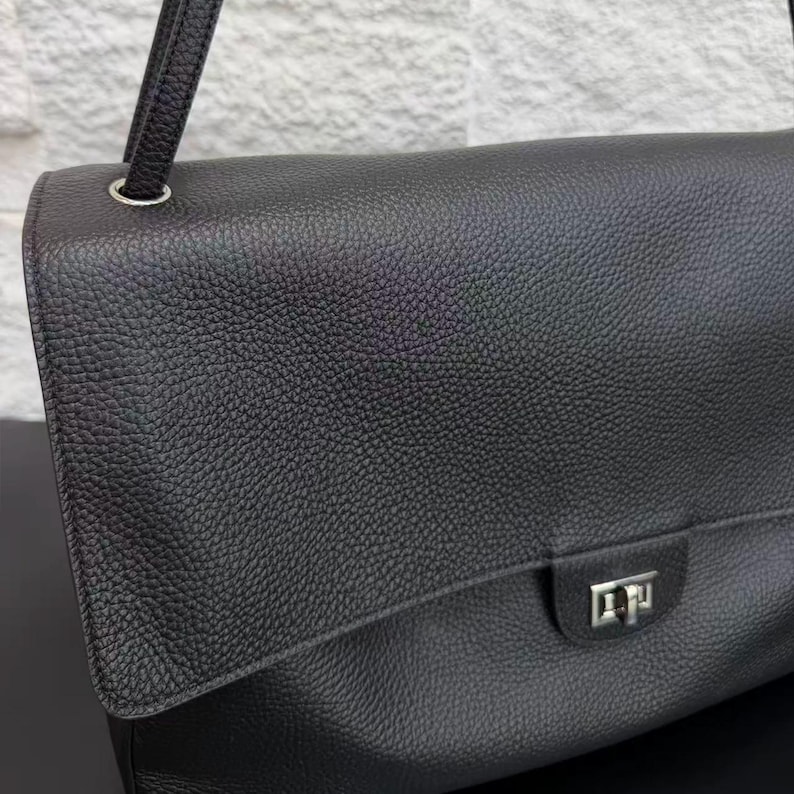 Detailed front view of a black leather shoulder bag featuring a textured leather flap, silver turn-lock closure, and adjustable strap attachment. The minimalist luxury design combines timeless style with practical functionality for everyday outfits and occasions.