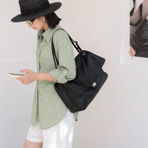 Woman wearing a black leather shoulder handbag with a classic flap design and silver buckle lock. The bag is styled as a chic everyday accessory with a comfortable shoulder strap, roomy interior, and elegant minimalist look suitable for casual outfits, office wear, and travel.