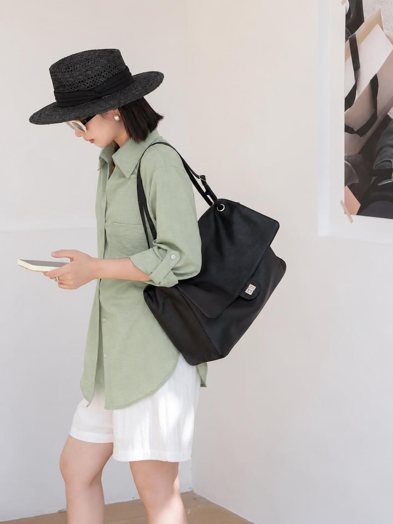 Woman wearing a black leather shoulder handbag with a classic flap design and silver buckle lock. The bag is styled as a chic everyday accessory with a comfortable shoulder strap, roomy interior, and elegant minimalist look suitable for casual outfits, office wear, and travel.